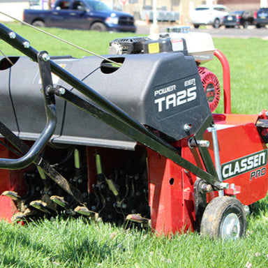 Classen-TA25DA-rear_view showing rear-side angle of TA25DA 25" PowerSteer PRO Aerator with visible tine cylinders engaging turf, black protective housings, red frame, and Honda GX120 engine, operating on green grass with urban vehicles in the background