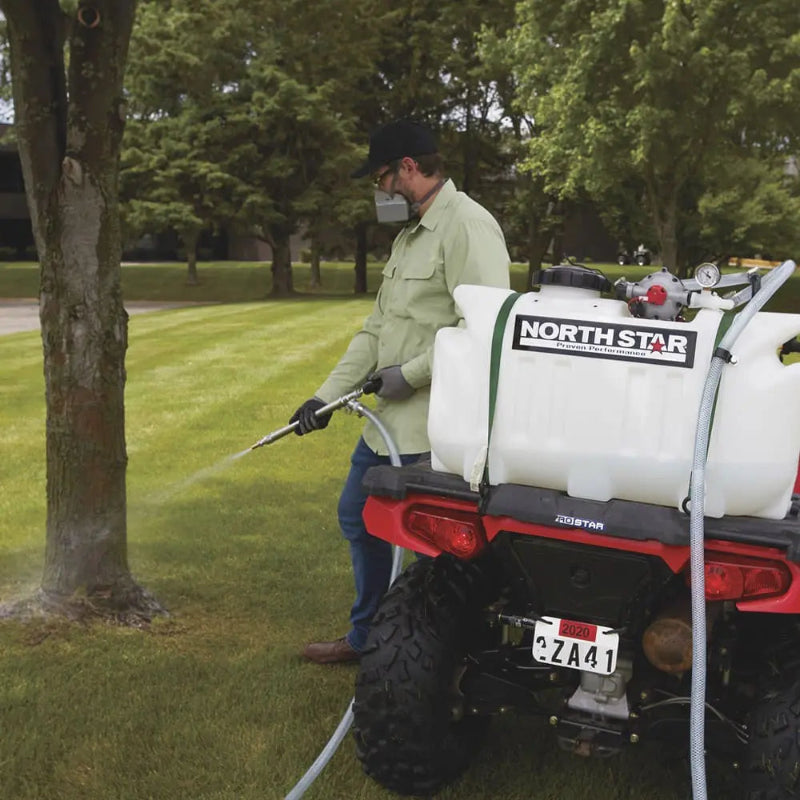 Man using NorthStar 12V ATV sprayer to water or apply herbicide around a tree, with the sprayer mounted on a red ATV and connected via a long hose for precise application.