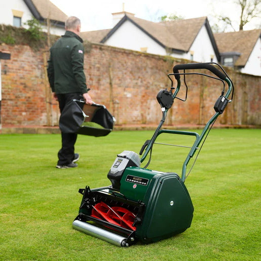 Allett Cambridge 17-inch battery-powered cylinder mower resting on a freshly cut lawn while a person walks away holding the grass catcher, showing its lightweight and convenient design.