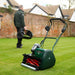 Allett Cambridge 17-inch battery-powered cylinder mower resting on a freshly cut lawn while a person walks away holding the grass catcher, showing its lightweight and convenient design.
