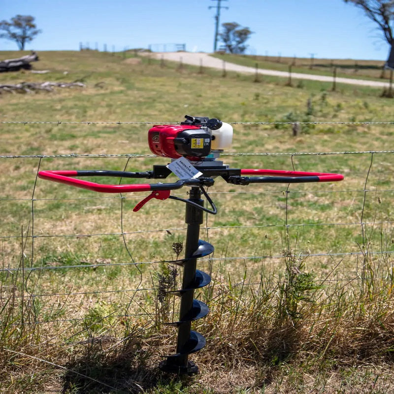 Bullmax BMEAFH-50HD earth auger in operation beside a wire fence in a rural field, with its red folding handles extended and auger bit engaged in the soil. Ideal for post-hole digging in agricultural or fencing installations.