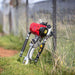 The image displays a Bullmax 4 Stroke Honda Post Driver, a gasoline-powered tool used for driving posts, leaning against a chain-link fence in a grassy outdoor setting. The tool features a red engine unit at the top and a silver, elongated body extending downwards.
