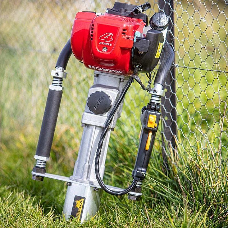 The image displays a Bullmax 4 Stroke Honda Post Driver, a gasoline-powered tool used for driving posts, leaning against a chain-link fence in a grassy outdoor setting. The tool features a red engine unit at the top and a silver, elongated body extending downwards.