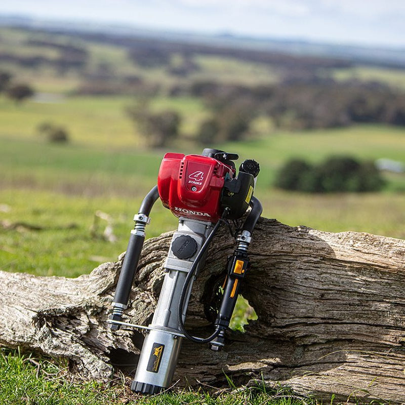 The image displays a Bullmax 4-Stroke Honda Post Driver, featuring a red engine and a silver-colored body, resting on a large, weathered piece of wood. The setting appears to be outdoors, with a blurred background of green hills and a clear sky, suggesting a rural or open landscape.