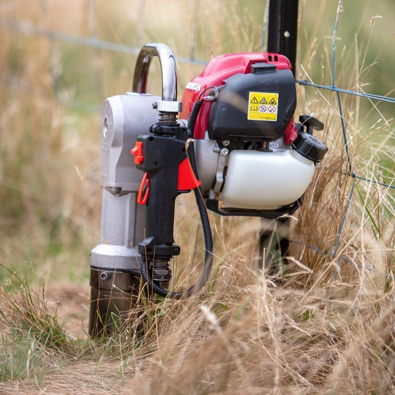 Bullmax BMPD-H80-2 post driver with Honda GX35 engine actively used in the field to drive a fence post into the ground. Surrounded by dry grass and wire fencing, the ergonomic grip and throttle are clearly visible during operation