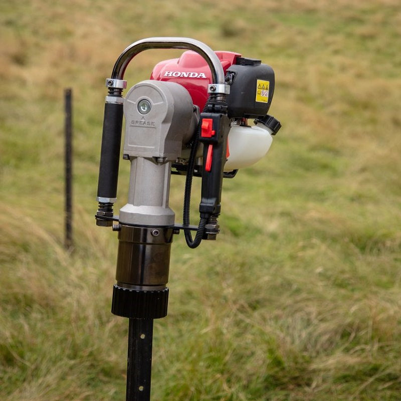 The Bullmax BMPD-H80-2 petrol post driver, featuring a Honda GX35 engine and dual ergonomic grips, is positioned vertically over a steel fence post in an open grassy field, demonstrating its efficiency in agricultural or rural fencing tasks