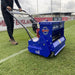 Side angle view of the Campey Turf Care Pedestrian AeraSeeder by Reist Industries being operated on a sports field, showing a worker pushing the blue overseeding machine with Honda GX engine, precision rollers, and seeding mechanism for turf maintenance and grass renovation.