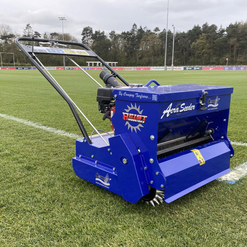 Side view of the Campey Turf Care Pedestrian AeraSeeder by Reist Industries in action on a sports field, featuring a Honda GX engine, spiked rollers for overseeding, and durable blue frame, designed for precise seeding and turf care maintenance.