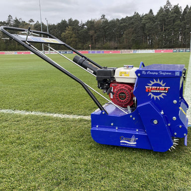 Side view of the Campey Turf Care Pedestrian AeraSeeder by Reist Industries in action on a sports field, featuring a Honda GX engine, spiked rollers for overseeding, and durable blue frame, designed for precise seeding and turf care maintenance.