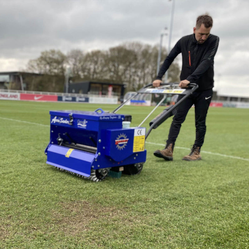 Rear angled view of the Campey Turf Care Pedestrian AeraSeeder by Reist Industries in operation, pushed by a groundskeeper on a sports field. The blue overseeding machine with Honda GX engine ensures precise turf renovation, overseeding, and aeration for professional grounds maintenance.