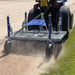 Close rear view of the Campey UniRake turf maintenance machine attached to a tractor, working on a sandy sports pitch. The spring tines are actively raking the surface, loosening soil, creating dust, and preparing the ground for professional turf conditioning.