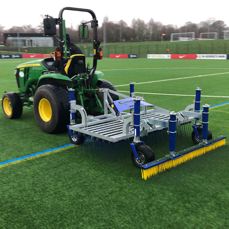 Campey UniRake attached to a John Deere tractor shown in action on an artificial sports pitch. The UniRake features spring tines and a rear brush, maintaining synthetic turf by lifting pile, removing debris, and evenly distributing infill across the surface.