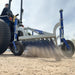 Low-angle action view of the Campey UniRake in operation, pulled by a tractor over a sandy surface. The spring tines are actively penetrating the ground, loosening material, improving aeration, and helping to maintain surface quality for sports turf and grounds.
