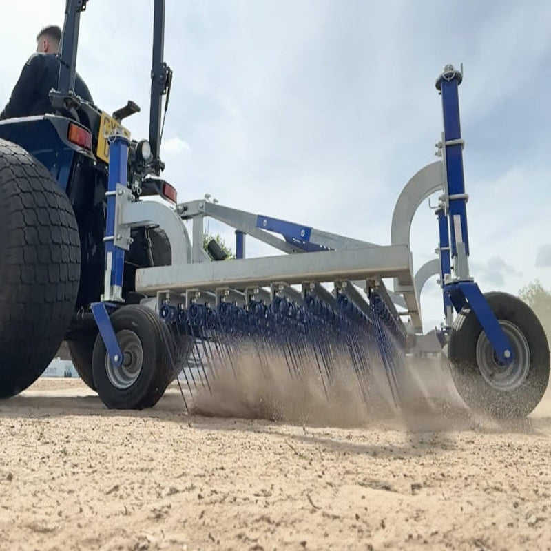 Low-angle action view of the Campey UniRake in operation, pulled by a tractor over a sandy surface. The spring tines are actively penetrating the ground, loosening material, improving aeration, and helping to maintain surface quality for sports turf and grounds.
