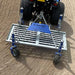 Overhead action view of the Campey UniRake connected to a tractor, working across a sandy surface. The steel frame with spring tines is clearly visible, raking and conditioning the ground to enhance aeration, leveling, and soil preparation for sports turf maintenance.
