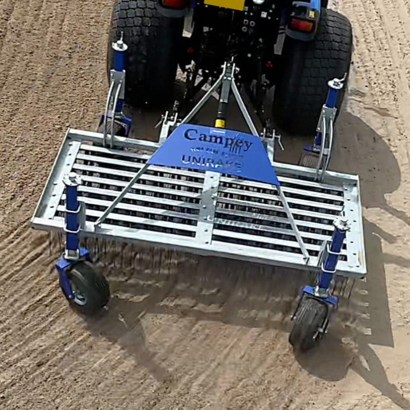 Overhead action view of the Campey UniRake connected to a tractor, working across a sandy surface. The steel frame with spring tines is clearly visible, raking and conditioning the ground to enhance aeration, leveling, and soil preparation for sports turf maintenance.