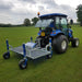 Rear field view of the Campey UniRake turf maintenance machine attached to a compact blue tractor. The UniRake’s spring tines work across a large grassy sports field, designed to aerate, decompact, and rejuvenate natural turf for improved playing performance.