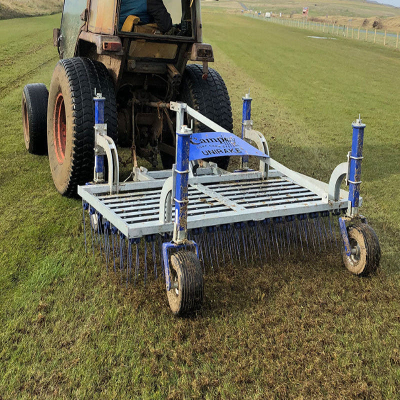 Campey UniRake attached to a tractor shown in rear view while working on a natural grass field. The UniRake’s spring tines penetrate and lift turf, improving aeration, removing organic debris, and enhancing field condition for better drainage and healthier playing surfaces.