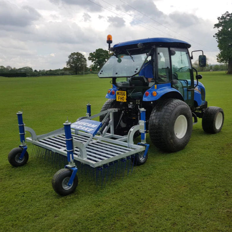 Rear field view of the Campey UniRake turf maintenance machine attached to a compact blue tractor. The UniRake’s spring tines work across a large grassy sports field, designed to aerate, decompact, and rejuvenate natural turf for improved playing performance.