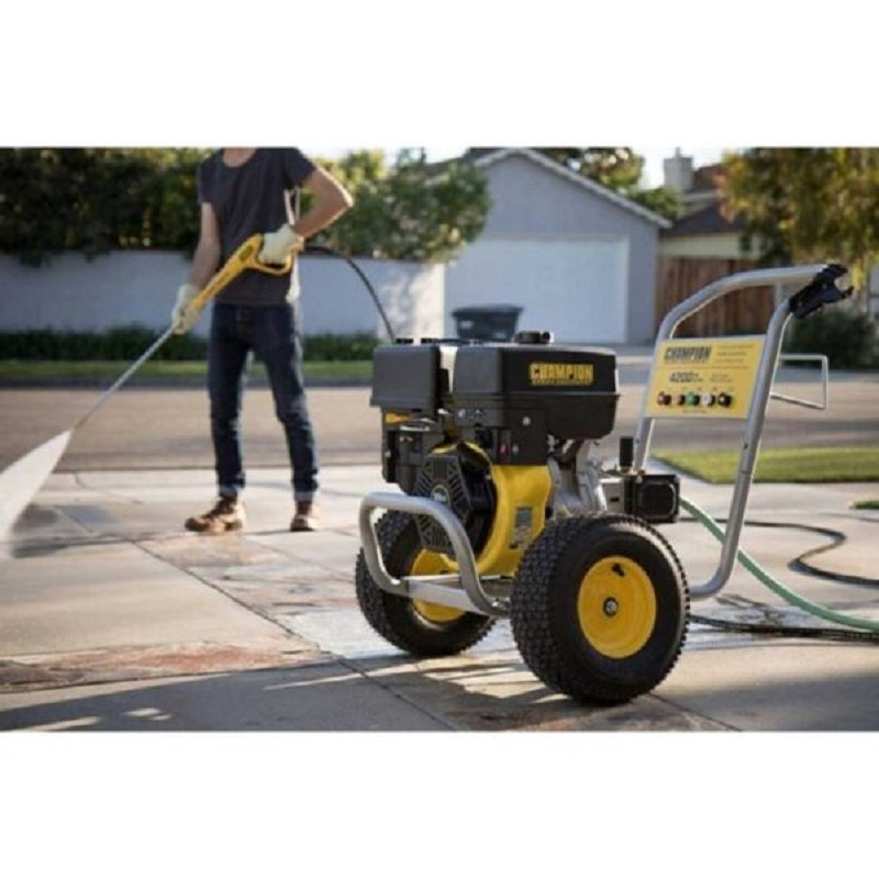 Person using the Champion 4200PSI Pressure Cleaner to wash a concrete driveway outdoors. The yellow and black unit is positioned on large wheels with the hose extended, spraying water powerfully, surrounded by houses and bright daylight.