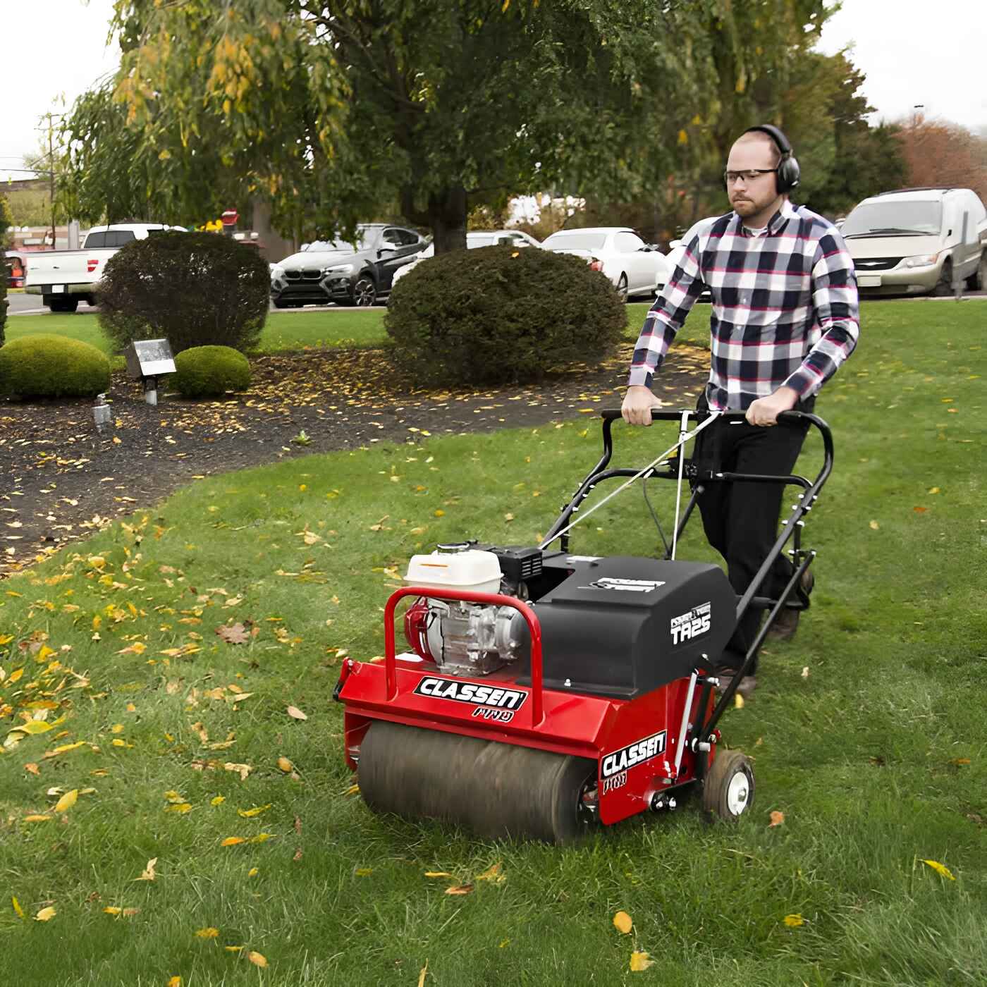A man operates the Classen PRO POWERSTEER Aerator TA-25DA on a fall-season lawn. Machine features red steel body, Honda engine, and ergonomic controls. Surrounding scene includes parked vehicles, trimmed bushes, and scattered leaves on lush green grass.