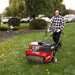 A man operates the Classen PRO POWERSTEER Aerator TA-25DA on a fall-season lawn. Machine features red steel body, Honda engine, and ergonomic controls. Surrounding scene includes parked vehicles, trimmed bushes, and scattered leaves on lush green grass.