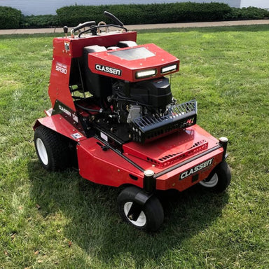Close-up view of the front LED light kit on the Classen SA-30 PRO aerator. Two rectangular LED lights are mounted on the red engine cover, providing enhanced visibility. The machine is positioned on a lawn with grass and sidewalk nearby.