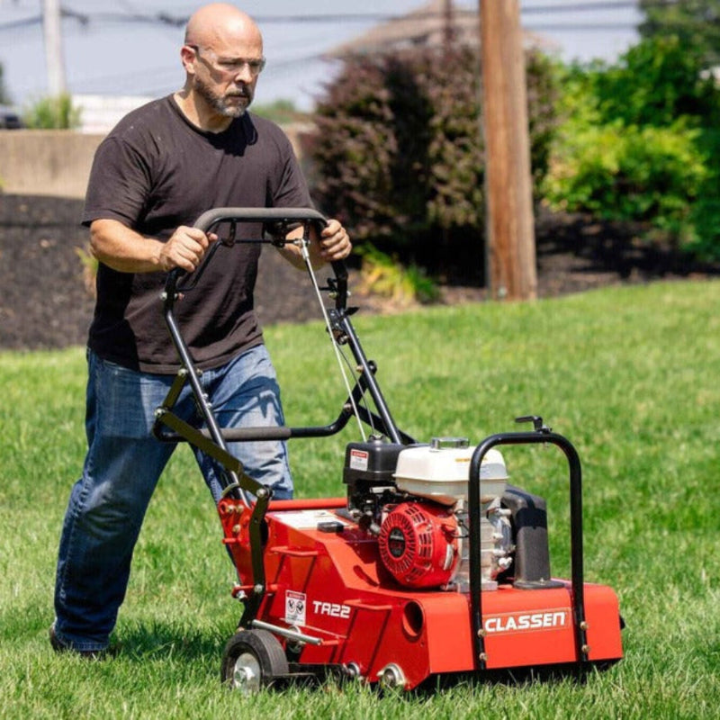 Classen_TA22H in action shows a man operating a red 21-inch steerable aerator with a Honda GX120 engine across a sunny green lawn. The aerator is in motion, with visible core tines, ergonomic handles, and residential landscaping in the background