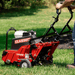 Classen TA22H 21 Aerator in action. shows a user guiding a red 21-inch steerable aerator with Honda GX120 engine across a grassy lawn. Soil plugs and turf debris are being ejected as the machine’s tines actively core and aerate the ground