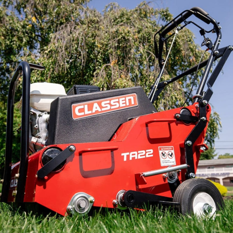 Side view of the Classen TA22H PRO Steerable Aerator on green grass, showcasing its red frame, compact design, control handle, black engine housing, and front caster wheel. Prominent Classen and TA22 labels highlight its identity as a turf aeration machine.