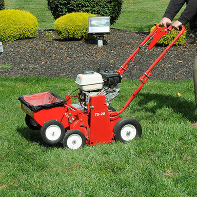 Person operating a red Classen TS-20 overseeder on a green lawn, surrounded by manicured bushes and mulch beds. The machine features a white fuel tank, black engine, dual wheels, and seed hopper, clearly labeled “TS-20,” in a landscaped outdoor setting.