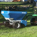 EcoLawn Applicator Eco 50 topdresser in action, attached to a John Deere tractor, spreading compost evenly across a grassy lawn. The blue hopper design with durable wheels ensures efficient, consistent soil and sand distribution for professional landscaping and turf maintenance.
