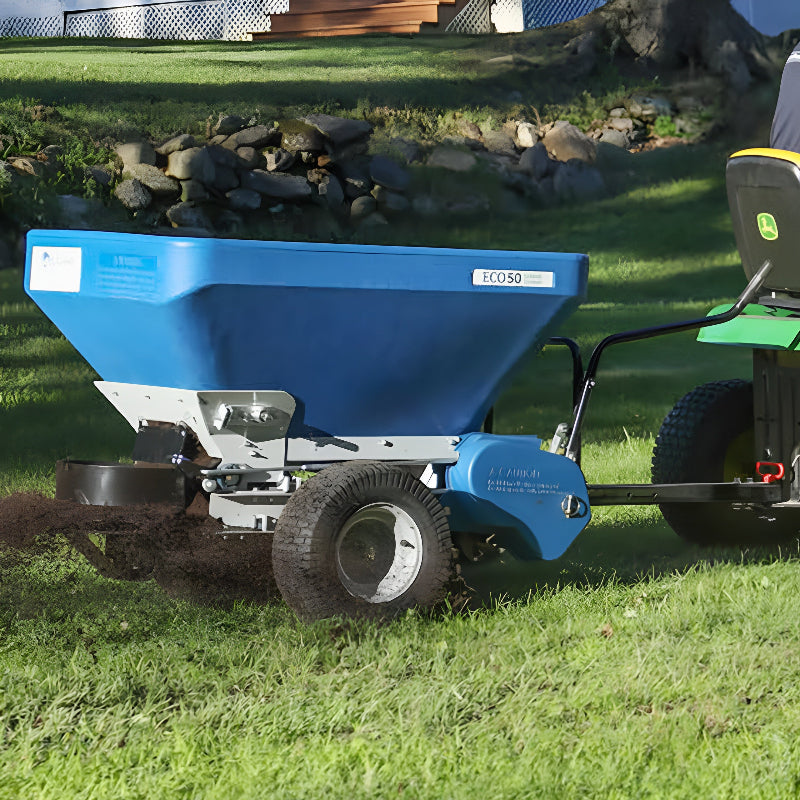 EcoLawn Applicator Eco 50 topdresser in action, attached to a John Deere tractor, spreading compost evenly across a grassy lawn. The blue hopper design with durable wheels ensures efficient, consistent soil and sand distribution for professional landscaping and turf maintenance.