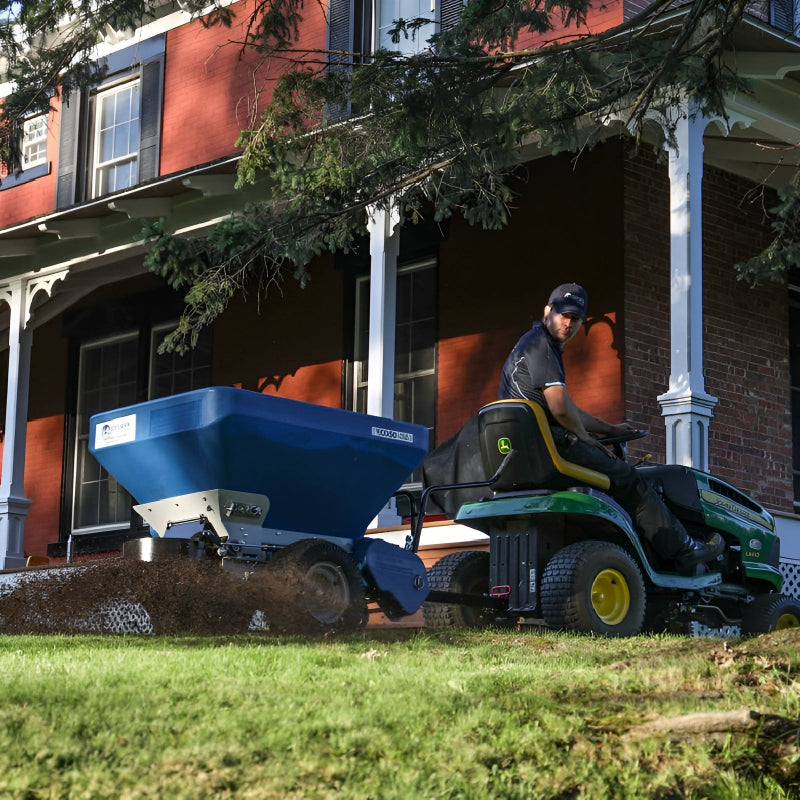 EcoLawn Applicator Eco 50 spreader attached to a John Deere tractor, spreading compost evenly on a residential lawn outside a red brick house. Operator ensures professional topdressing for turf health, soil improvement, and sustainable landscape maintenance in a controlled environment.