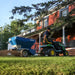 EcoLawn Applicator Eco 50 spreader attached to a John Deere lawn tractor, distributing topdressing material evenly on a residential lawn. Operator in action near a red brick house ensures smooth turf care, efficient soil spreading, and professional landscaping results.