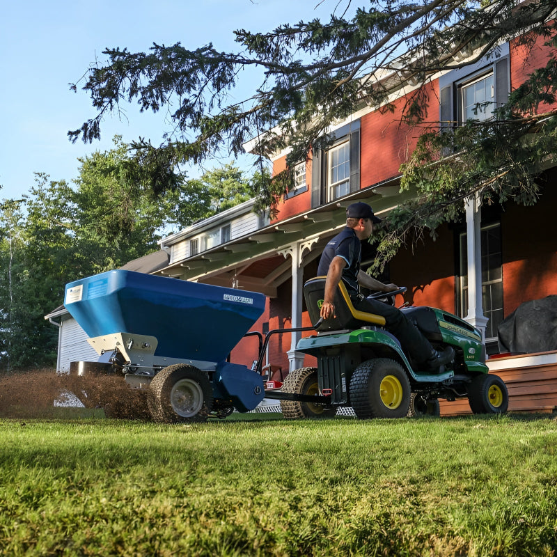 EcoLawn Applicator Eco 50 spreader attached to a John Deere lawn tractor, distributing topdressing material evenly on a residential lawn. Operator in action near a red brick house ensures smooth turf care, efficient soil spreading, and professional landscaping results.