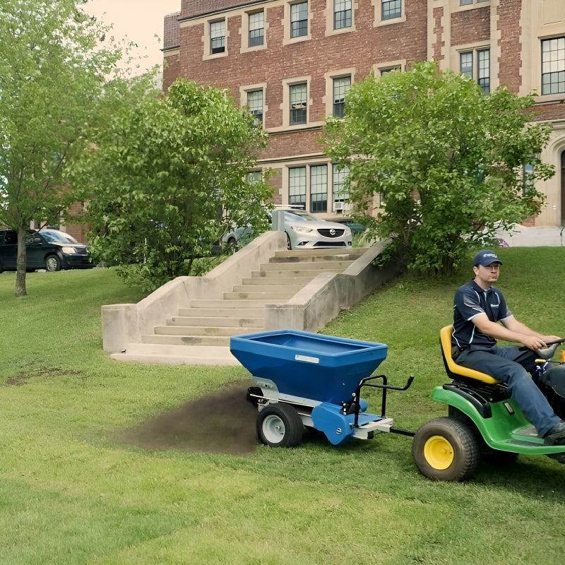 EcoLawn Applicator Eco 50 tow-behind spreader in action, pulled by a John Deere tractor, applying compost topdressing on a green lawn outside a historic brick building. Operator ensures precise distribution for turf renovation, soil health, and professional landscape maintenance.