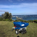 Rear view of the Eco 250 Top Dressing Spreader on a grassy coastal hillside, facing the ocean. The blue hopper and robust tires stand out against the scenic seascape, showcasing its reliability for turf care in diverse outdoor environments.