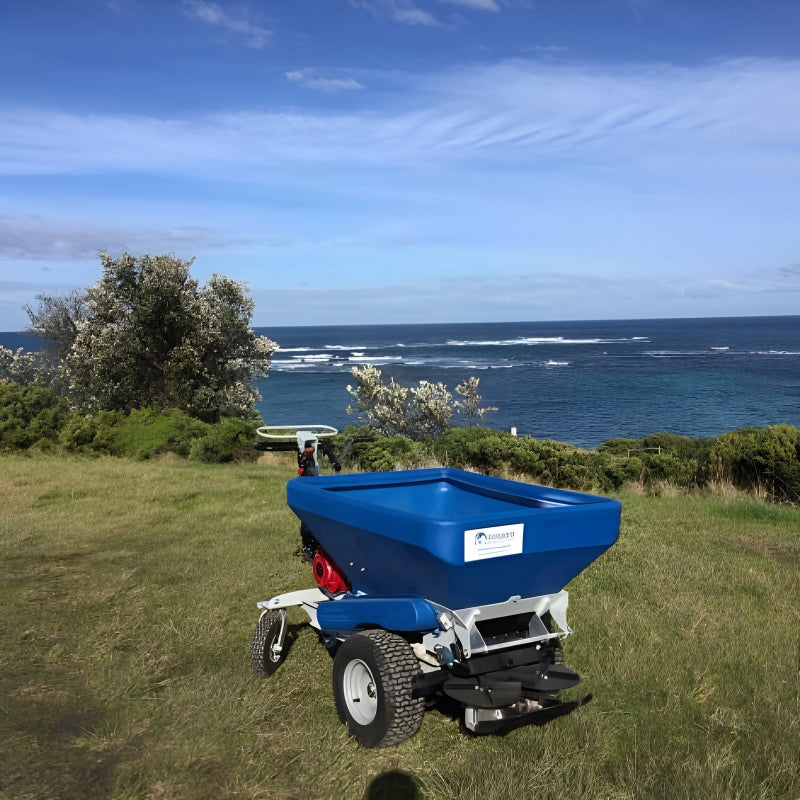 Rear view of the Eco 250 Top Dressing Spreader on a grassy coastal hillside, facing the ocean. The blue hopper and robust tires stand out against the scenic seascape, showcasing its reliability for turf care in diverse outdoor environments.