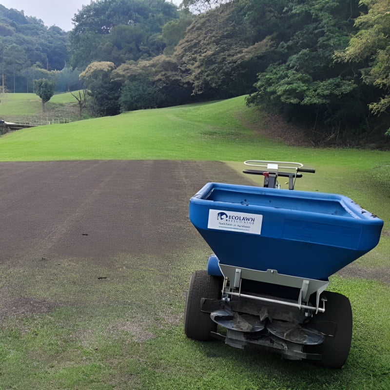 Rear view of the Eco 250 Top Dressing Spreader operating on a golf course lawn. The blue hopper and spreading system are visible as it applies topdressing material, surrounded by lush green grass and trees, demonstrating professional turf care efficiency.