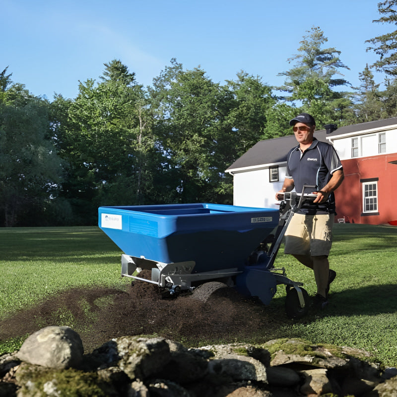 A lawn care professional uses the Eco 250 Top Dressing Spreader to evenly distribute soil across a well-maintained residential lawn. The machine’s blue hopper and spreading mechanism are clearly visible as it operates efficiently near a stone border and house.
