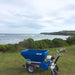 Side view of the Eco 250 Top Dressing Spreader positioned on grassy terrain overlooking the ocean. The blue hopper and sturdy wheels highlight its build, while the scenic coastal backdrop emphasizes its use in diverse outdoor turf maintenance environments.