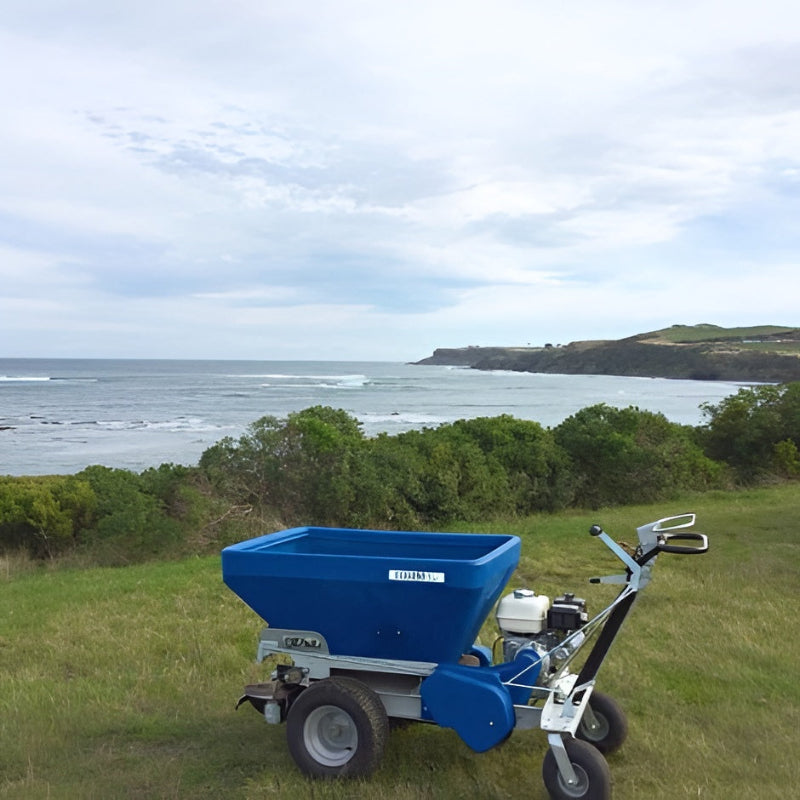 Side view of the Eco 250 Top Dressing Spreader positioned on grassy terrain overlooking the ocean. The blue hopper and sturdy wheels highlight its build, while the scenic coastal backdrop emphasizes its use in diverse outdoor turf maintenance environments.