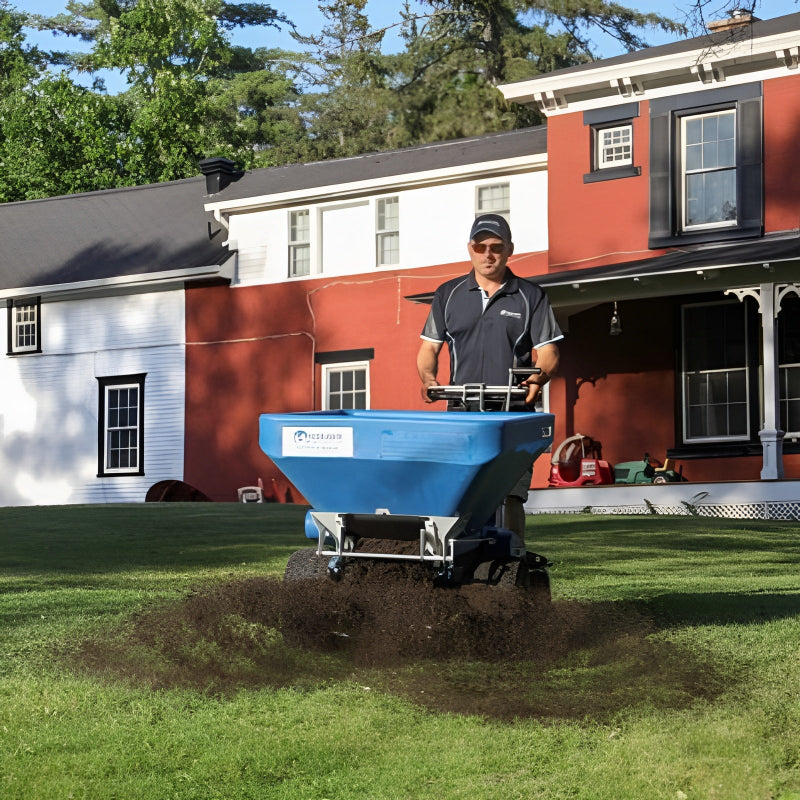 A man uses the Eco 250 Top Dresser to spread soil evenly across a green residential lawn. The blue machine applies topdressing in front of a red and white house, improving turf health and ensuring professional lawn care results.