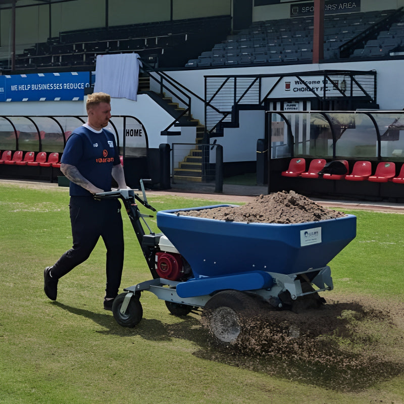 A sports field worker pushes the Eco 250 Top Dresser filled with soil across a grass pitch. The machine distributes topdressing evenly while stadium seating and dugouts are visible in the background, showcasing turf maintenance during field renovation work.