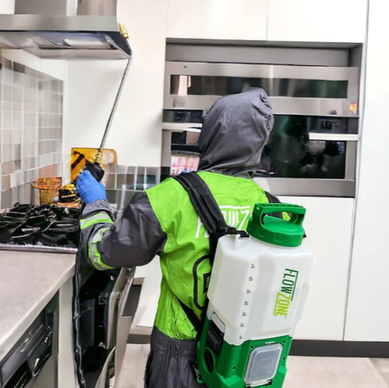 The image depicts an individual wearing protective gear, including a hooded suit and gloves, using a FlowZone backpack sprayer inside a kitchen. The person is positioned near a stove and kitchen cabinets, operating the sprayer, which is designed for various applications like pest control or disinfection. The sprayer itself is a prominent feature, labeled "FLOWZONE" on its side. 