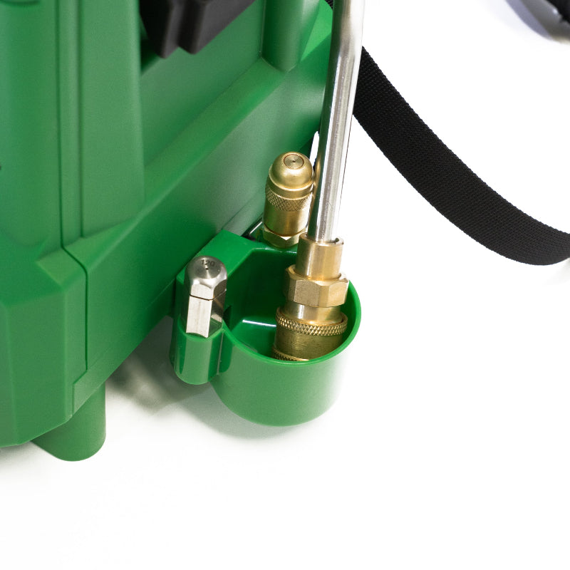 The image shows a close-up view of a green component of what appears to be a sprayer unit, specifically identified as part of a FlowZone Typhoon G3 + Swap Tank MicroBurst Pest Pack. The green component features a molded holder designed to secure a metallic spray wand, which is partially visible. Brass fittings are prominently displayed, including a valve mechanism and connection points for the wand and potentially other components of the spraying system. A black strap, likely part of a shoulder harness for 