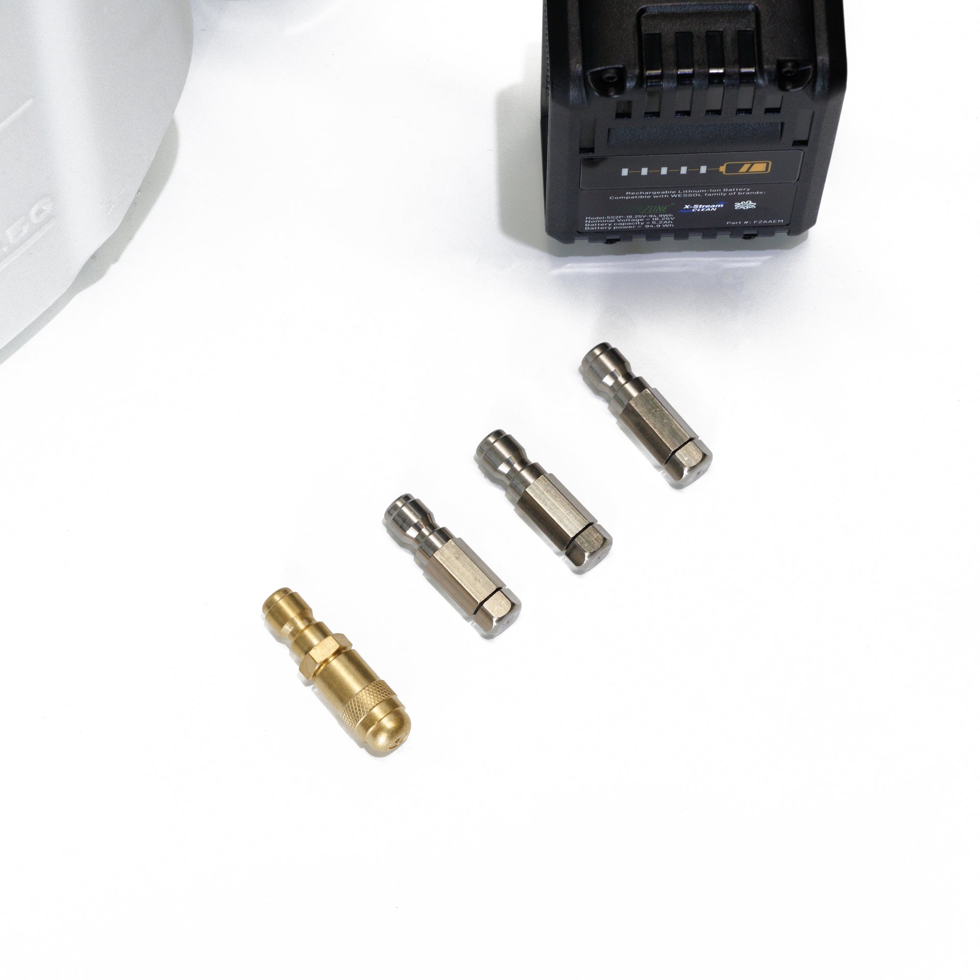 Close-up view of FlowZone MicroBurst 10L accessories displayed on a white surface, featuring one brass nozzle and three stainless steel nozzles neatly lined up. The edge of the battery pack and sprayer tank are partially visible in the frame.
