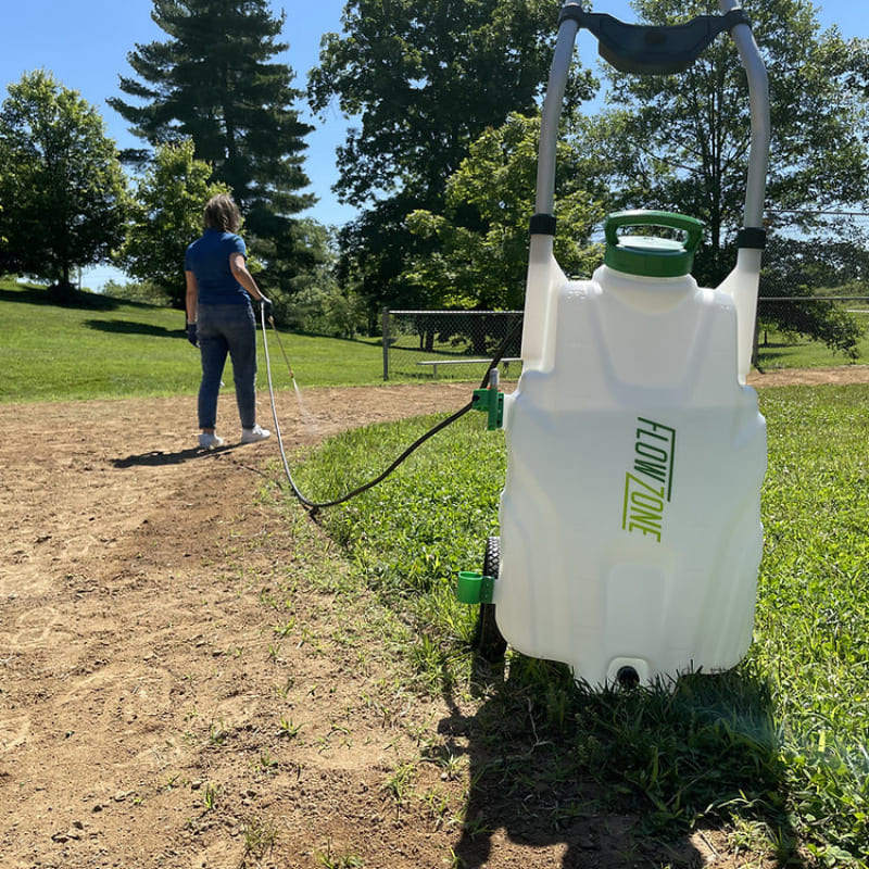 FlowZone Monsoon 2.5 34L battery trolley sprayer in use outdoors, positioned on grass with spray wand extended by a person treating soil, photographed from a low rear-side angle under sunny conditions with trees and blue sky in background.