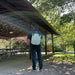 Rear outdoor view of a person wearing a FlowZone Storm 2.5 10L battery-powered backpack sprayer, spraying under a wooden pavilion roof. The white tank with green lid and black straps is clearly visible, highlighting portability and hands-free spraying convenience.